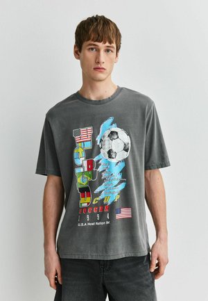 Mladý muž nosiaci sivé tričko z majstrovstiev sveta v futbale USA 1994 s veľkým obrázkom futbalovej lopty a vlajkami vpredu.