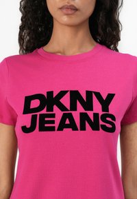 Vaaleanpunainen puuvillapaita, jossa on suuri musta "DKNY JEANS" -logo edessä. Pyöreä kaula-aukko ja lyhyet hihat. Sujuva rakenne.