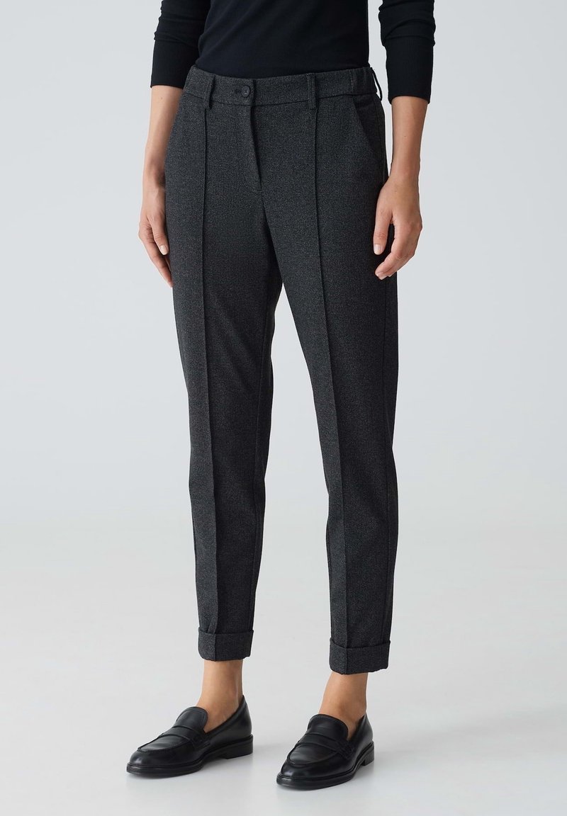 Femme portant un pantalon ajusté gris foncé avec des ourlets retournés et des mocassins en cuir noir, debout devant un fond clair uni.