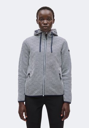 Femme portant une veste à capuche zippée à motif avec cordons et un pantalon noir, debout face à l’avant avec une expression neutre.