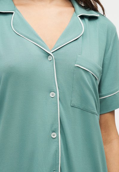 Chemise vert clair à manches courtes avec boutons, avec passepoil blanc sur le col, la poche et les poignets, portée par une personne aux cheveux foncés.