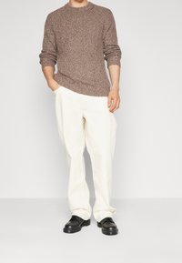 Brązowy sweter dzianinowy z okrągłym dekoltem, połączony z białymi spodniami i czarnymi butami. Sweter ma teksturowany, gruby wzór.