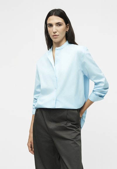 Γυναίκα με μακριά σκούρα μαλλιά που φοράει ένα ανοιχτό μπλε πουκάμισο με στάντ γιακά και μαύρο παντελόνι με ψηλή μέση, στέκεται με το ένα χέρι στην τσέπη.