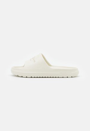 Hvid slip-on sandal med tyk sål og en bred rem prydet med et mærkelogo på siden.