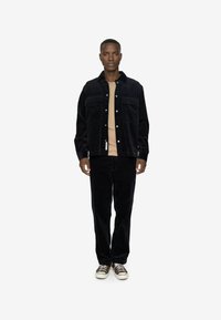 Black corduroy jacket with two chest pockets, paired with matching corduroy pants. Underneath, a light beige shirt. Casual sneakers complete the look.