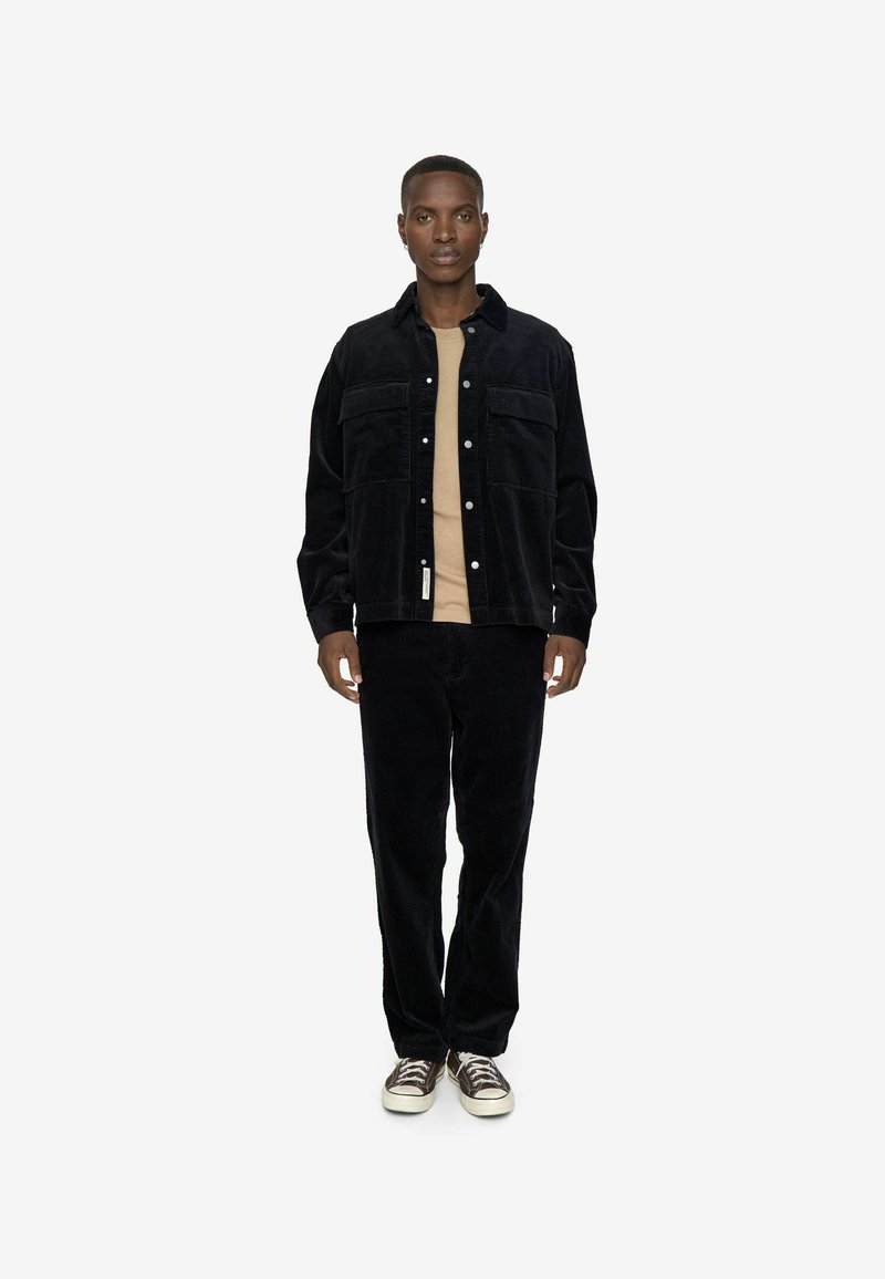 Black corduroy jacket with two chest pockets, paired with matching corduroy pants. Underneath, a light beige shirt. Casual sneakers complete the look.