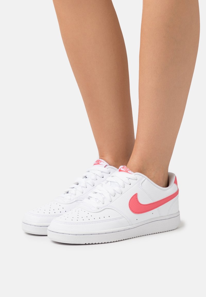 Baskets en cuir blanc avec des accents roses, présentant des perforations sur le bout et une semelle texturée. Les lacets sont blancs avec le logo de la marque.