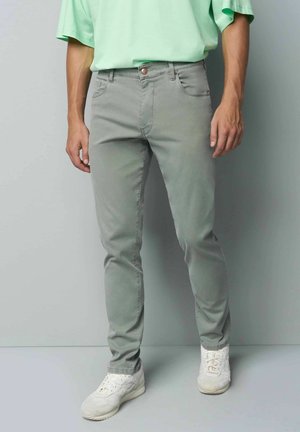 Mann trägt hellgraue Slim-Fit-Jeans, weiße Turnschuhe und ein hellgrünes kurzärmeliges Hemd, steht vor einer schlichten Wand.