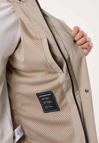 Beige jakke åbnet for at vise åndbar, perforeret inderfor med mærke for vandtæt, åndbar og vindtæt samt inderlomme.