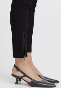 Crne krojene hlače s patentnim zatvaračima na zapešćima, u kombinaciji s crnim sandalama na petu s izrezima i blok petom.