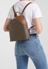 Brown leather backpack with a patterned lower section, curved top, and dual shoulder straps; zipper closure and top handle for carrying.