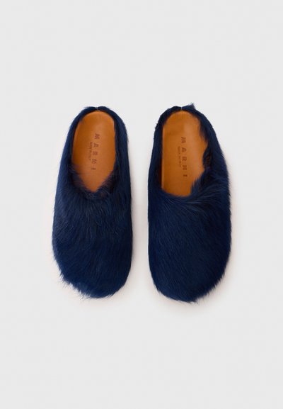Chaussons en fourrure bleu marine avec une texture douce, une forme arrondie et un design flexible. Doublure en cuir à l'intérieur avec le nom de la marque embossé.