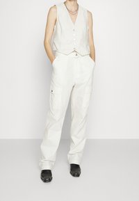 Vit ärmlös väst med knappar som kombineras med lösa vita cargobyxor med sidofickor och svarta anklarstövlar.