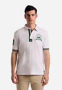 Camisa polo blanca con detalles verdes. Presenta una tapeta de tres botones, un logo bordado en el pecho y puños a rayas. Mangas cortas.