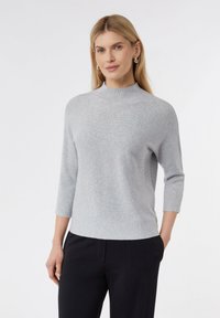 Lichtgrijze, geribbelde gebreide trui met een hoge hals en driekwartmouwen, gecombineerd met donkere broek. Eenvoudig ontwerp, zachte textuur.