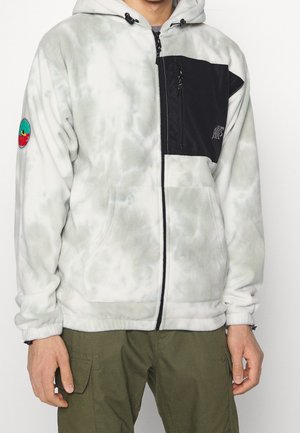 Mann trägt weißen und grauen Batik-Fleece-Hoodie mit Reißverschluss, schwarzer Brusttasche und grüner Cargo-Hose, steht vor einem schlichten Hintergrund.