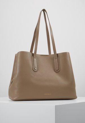 Torba typu tote z naturalnej skóry w kolorze taupe z długimi podwójnymi uchwytami, subtelną fakturą i małym złotym logo marki z przodu, wyeksponowana na białej powierzchni.