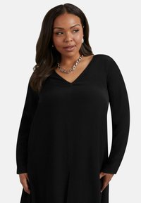 Svart långärmad klänning med V-ringning. Har en mjuk textur och en avslappnad silhuett. Silver halsband ger kontrast.