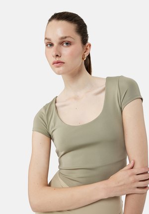 Ung kvinde med trukket hår tilbage iført små gyldne øreringe i ringform og en tætsiddende, lys olivengrøn kortærmet top mod en ensfarvet baggrund.