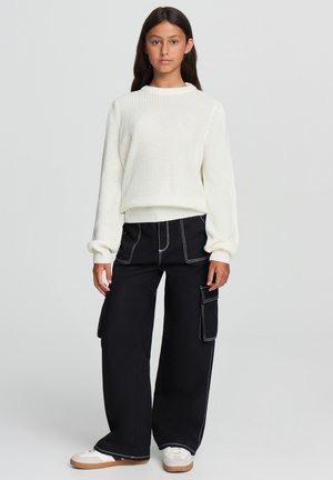 Maglione bianco a coste con maniche lunghe abbinato a pantaloni cargo neri a gamba larga con tasche laterali e cuciture bianche, indossato con sneakers bianche.