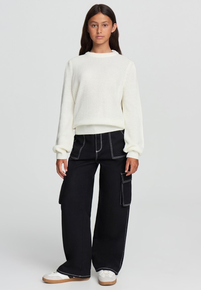 Biały sweter z prążkowanego dzianiny z długimi rękawami, w połączeniu z czarnymi szerokimi spodniami cargo z kieszeniami bocznymi i białymi przeszyciami, noszony z białymi trampkami.