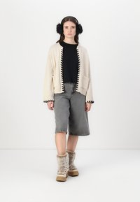 Cremefarbener Cardigan mit schwarzem Saum, schwarzes Hemd, graue Jeansshorts und flauschige beigefarbene Stiefel. Der Cardigan hat zwei Fronttaschen.
