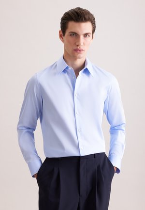 Ung mand iført en lyseblå skjorte med knapper og krave samt sorte bukser, står med hænderne i lommerne foran en ensfarvet baggrund.