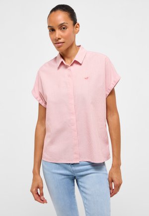 Woman wearing a short-sleeve, light pink button-up shirt with small stripes and light blue jeans, standing against a plain background.