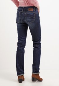 Tmavě modré skinny džíny s mírným vyblednutím, zadní kapsy se švy a hnědá kožená nášivka na pase. Nosené s béžovými kozačkami do kotníků.