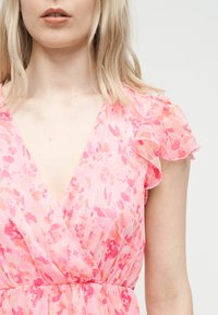 Ξανθιά γυναίκα που φοράει ροζ και κοραλλί φόρεμα τύπου wrap με κοντά ριγέ μανίκια, σε απλό λευκό φόντο.