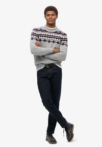 Grijze gebreide trui met een patroon op de schouders, donkere spijkerbroek en zwarte veterschoenen. Model poseert met de armen gekruist om de details van de outfit te tonen.