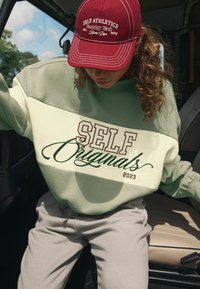 Groene en crèmekleurige sweatshirt met "SELF Originals 2023" in contrasterende letters. Bordeau-rode pet met geborduurde tekst. Grijze broek.