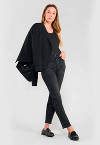 Blazer negro ajustado colocado sobre el hombro, encima de una camiseta sin mangas negra, combinado con vaqueros negros de talle alto y mocasines negros brillantes.