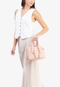 Borsa a tracolla rosa strutturata con una superficie liscia, doppio manico superiore e dettagli in catena dorata, tenuta da una persona che indossa un gilet bianco e una gonna beige.