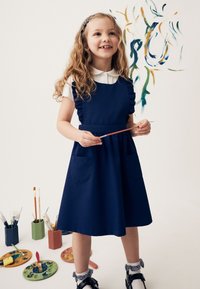 Jeune fille heureuse en robe bleu marine tenant un pinceau avec des fournitures de peinture et des œuvres d'art colorées sur un mur blanc derrière elle.