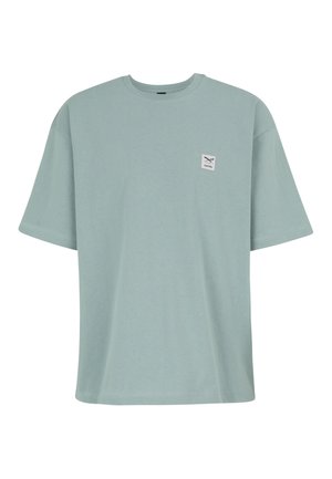 Hellblaues oversized T-Shirt mit kurzen Ärmeln, Rundhalsausschnitt und kleinem weißem quadratischem Aufnäher mit gekreuzten Werkzeugen auf der linken Brust.