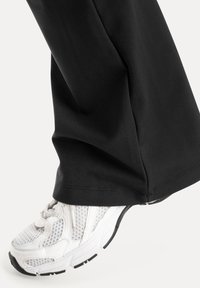 Chaussure de course en mesh blanc portée avec un pantalon noir, montrant la partie inférieure de la jambe et le pied en position légèrement relevée.