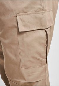 Pantaloni cargo bej, cu un buzunar lateral cu capac, confecționați dintr-un material neted și durabil, cu un finisaj curat.