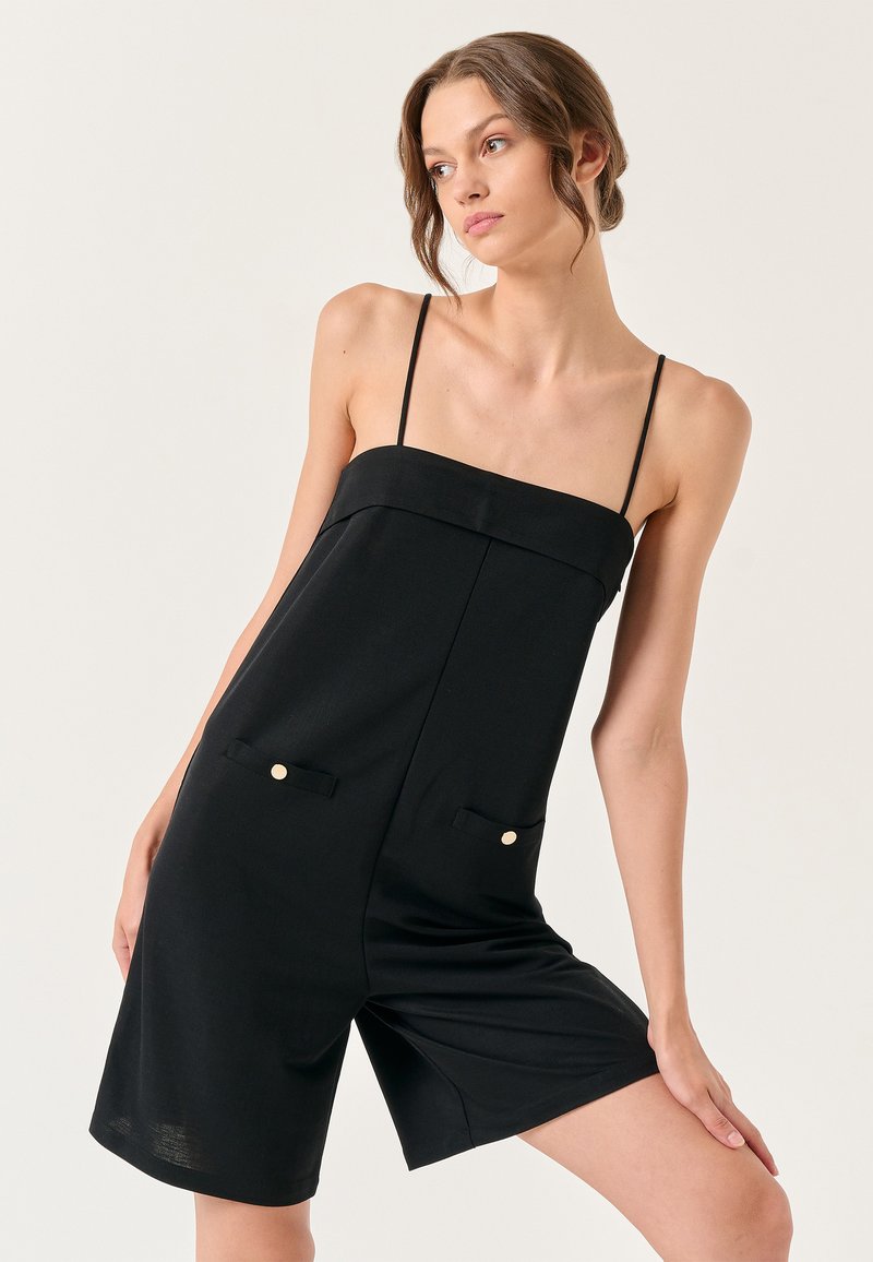 Zwarte playsuit met dunne bandjes, vierkante halslijn, voorzakken en gouden knoopaccenten. Glad materiaal met een strakke pasvorm en relaxte shorts.