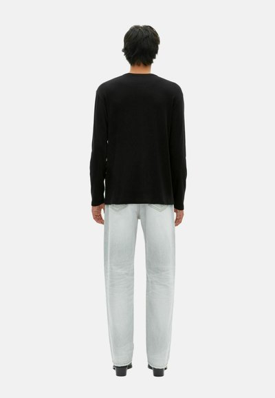 Personne se tenant de dos, portant un t-shirt à manches longues noir, un jean bleu clair et des chaussures noires, sur un fond uni clair.