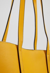 Sac à main en cuir jaune avec des anses bordées de noir et des coutures visibles sur un fond clair uni.