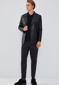 Zwarte leren jas met gestructureerde revers en een voorzak, gecombineerd met stijlvolle grijze textuurbroeken en zwarte schoenen. Over het geheel een gladde textuur.