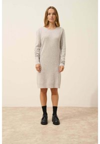 Abito in maglia grigia chiara con maniche lunghe e texture a costine, di lunghezza al ginocchio, abbinato a scarpe e calze nere su uno sfondo neutro.