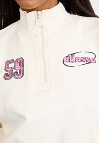 Felpa bianca con zip e colletto alto. Presenta il numero ricamato "59" in rosa e il logo "ellesse" con contorno blu. Texture morbida.