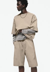Top bej oversized cu maneci scurte, stratificat peste o cămașă lungă gri deschis, asociat cu pantaloni scurți bej cu șnur asortat. Detalii subtile de cusătură.