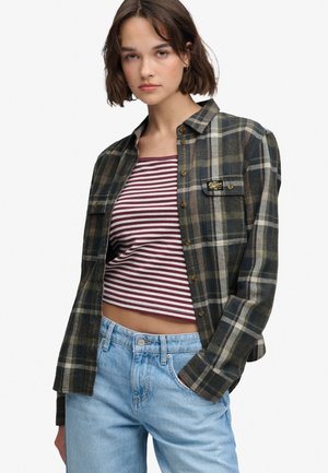 Ung kvinde med kort brunt hår iført en grønt ternet skjorte med knapper over en rød og hvid stribet crop top og lyseblå jeans.