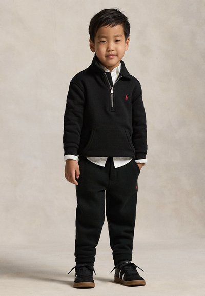 Jeune garçon debout portant un sweat noir à fermeture éclair, un pantalon noir, une chemise blanche en dessous et des baskets noires avec des semelles en caoutchouc.