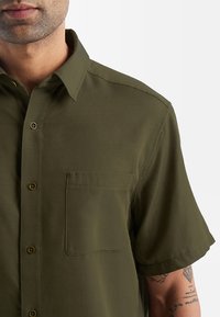 Olijfgroen overhemd met korte mouwen, gemaakt van zachte stof, met een button-down kraag, een borstzak en bruine knopen.
