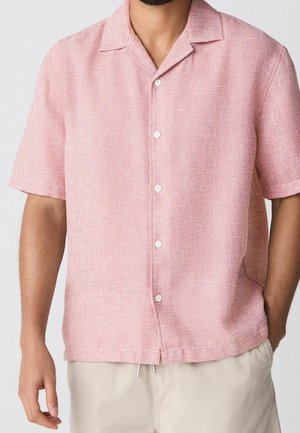 Homme portant une chemise texturée rose clair à manches courtes avec boutons et un pantalon beige clair, la main droite dans la poche.