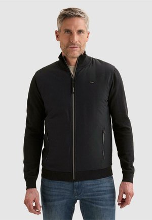 Mann trägt schwarze Jacke mit Reißverschluss, geripptem Kragen, gerippten Bündchen und zwei vorderen Reißverschlusstaschen, kombiniert mit blauen Jeans, vor einfarbigem Hintergrund.